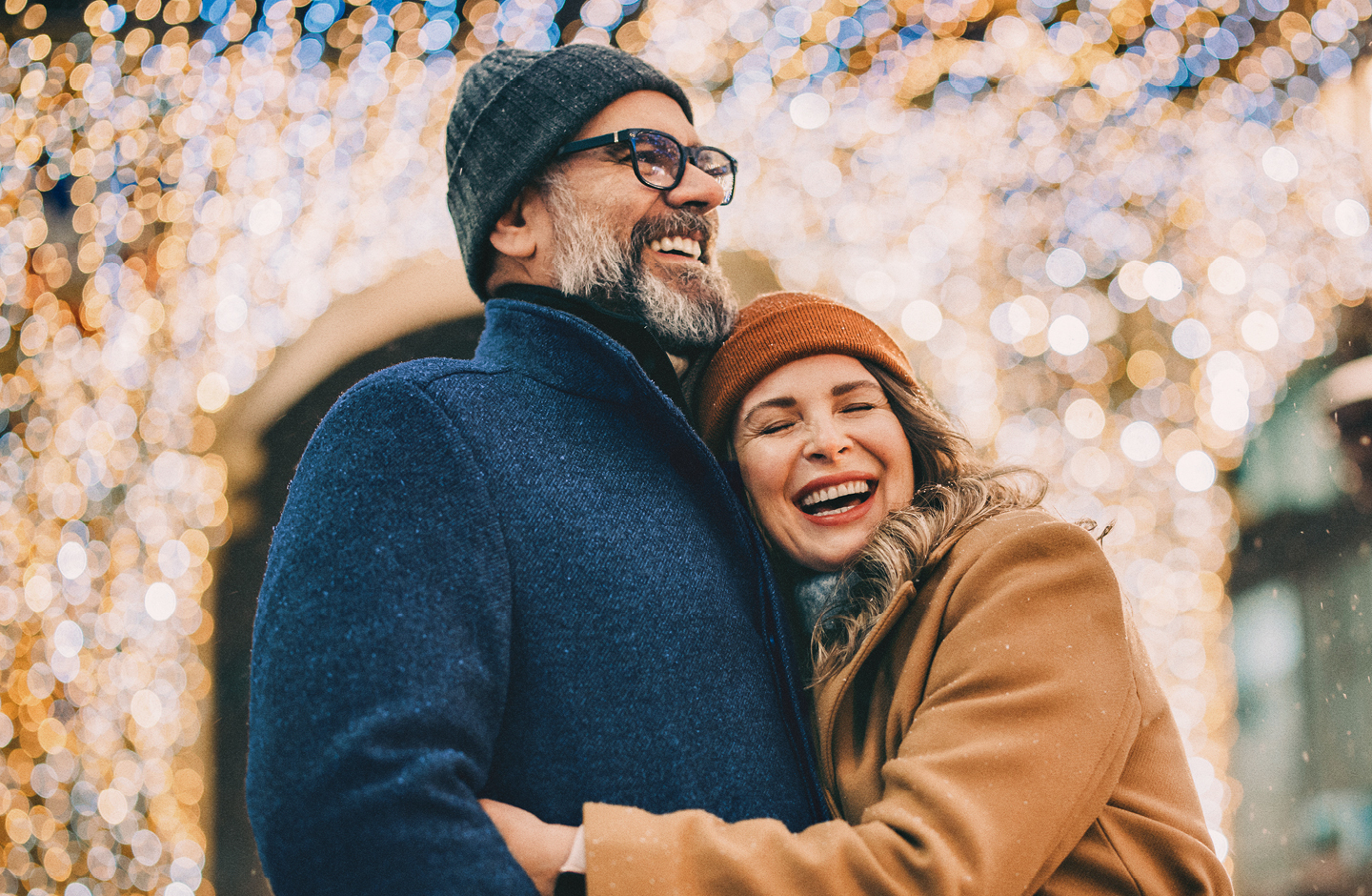 Ein älteres Paar steht vor einem glitzernden Hintergrund und umarmt sich. Beide tragen Winterkleidung und lächeln.