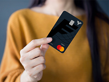 Eine Frau in einem gelben Pullover hält die Volksbank Debit Mastercard Business in der Hand. Man kann ihr Gesicht nicht sehen.