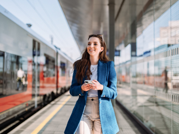 Eine junge Frau in blauem Blazer steht auf einem Bahngleis mit Handy in der Hand und lacht.