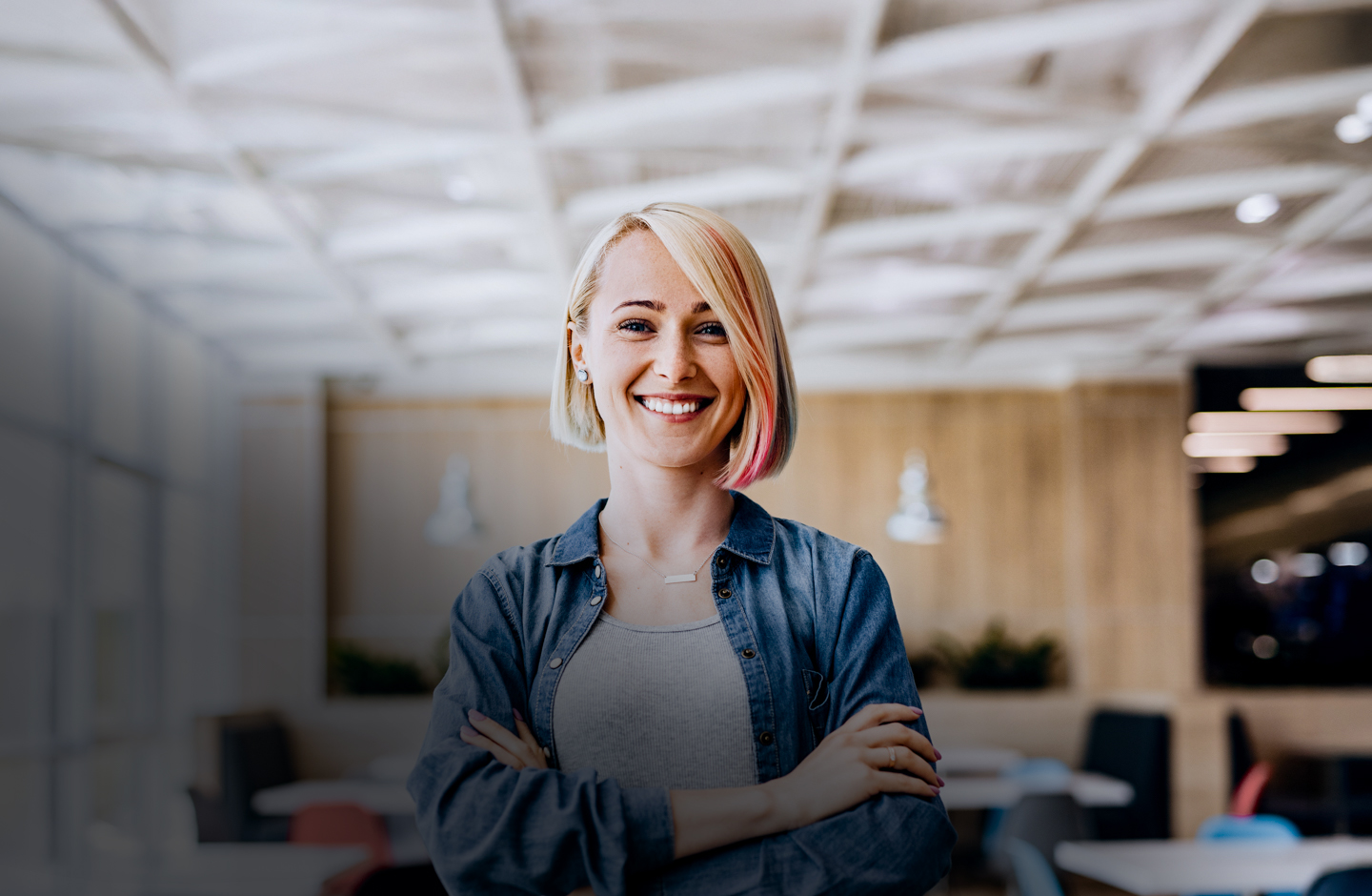 Eine Frau mit kurzen, blonden Haar verschränkt in einem Büro lachend die Arme.