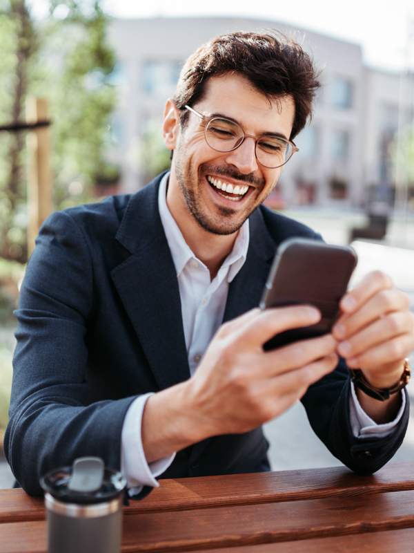Ein Mann sitzt im Freien auf einer Bank und Blickt mit einem breiten Lächeln auf sein Mobiltelefon.